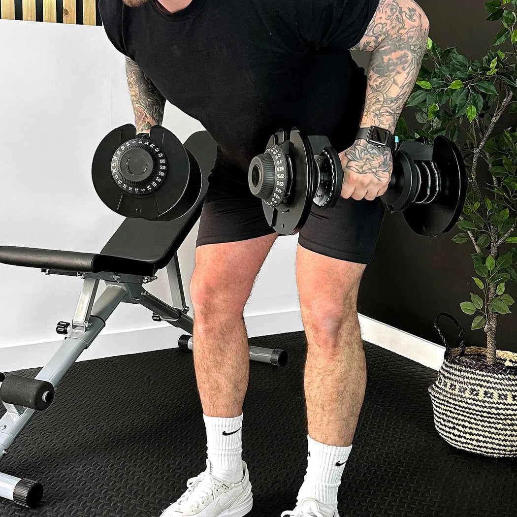 Person lifting dumbbells in a gym setting with a bench and plant in the background