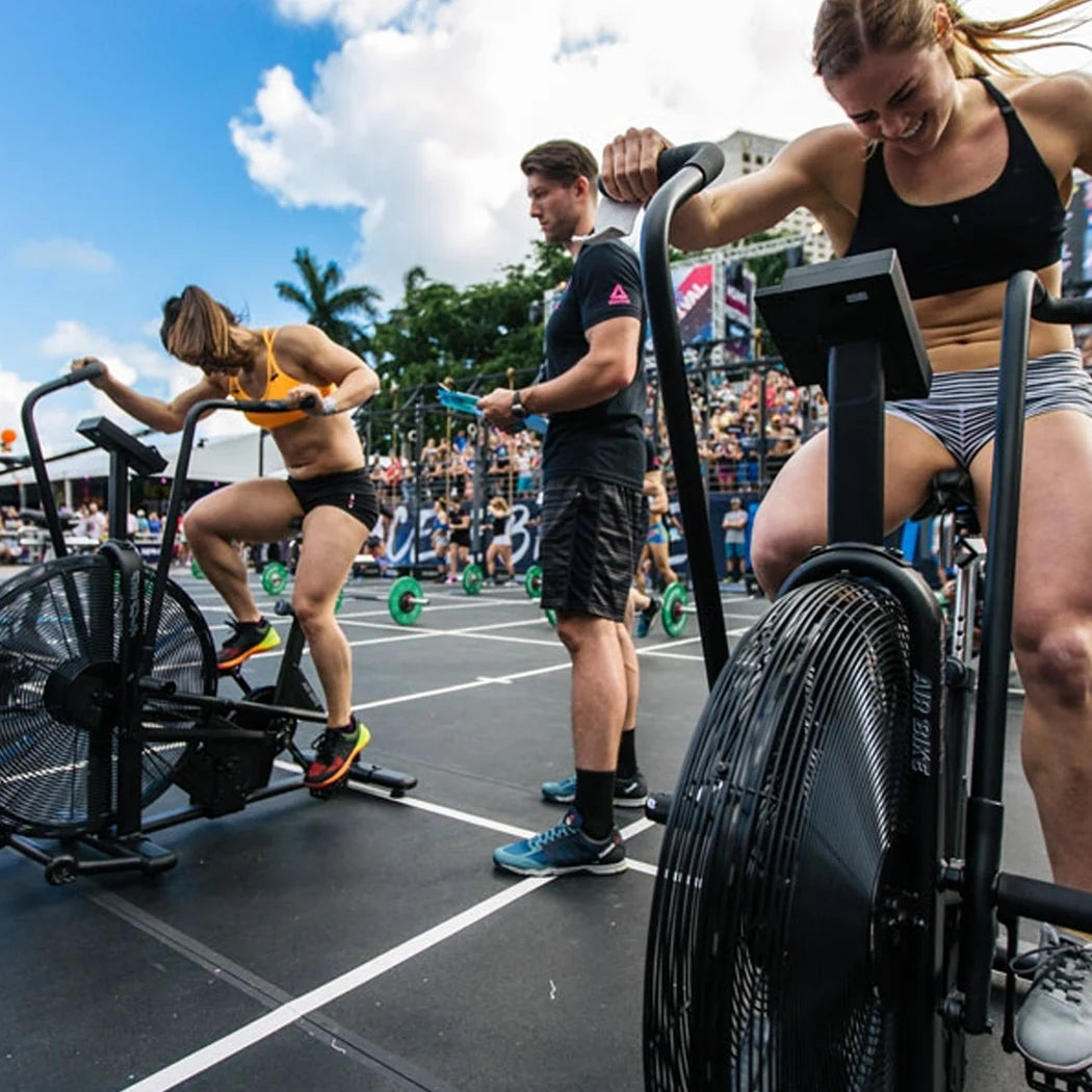 Two people using exercise bikes outdoors with a crowd and buildings in the background