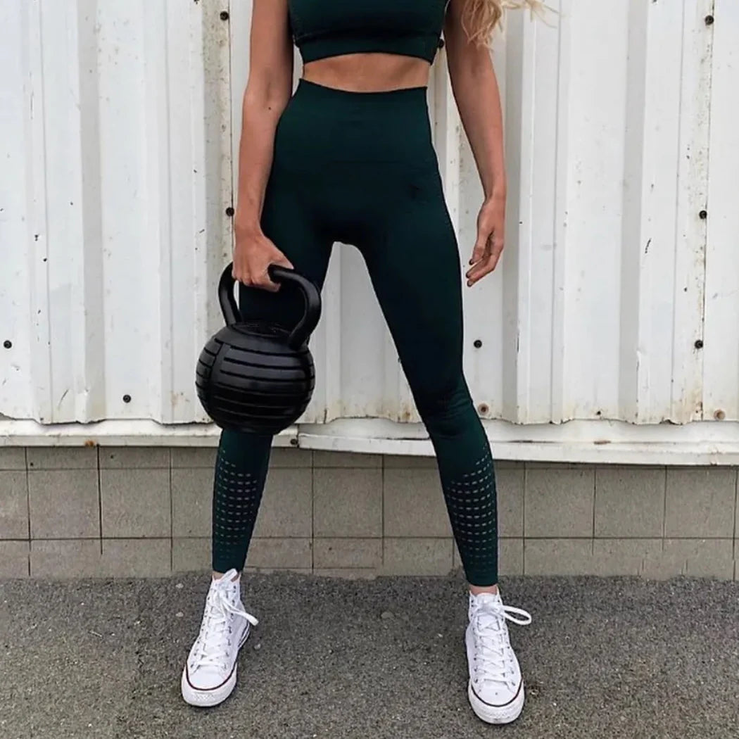 Person holding a black kettlebell wearing dark green leggings and white sneakers against a white wall.