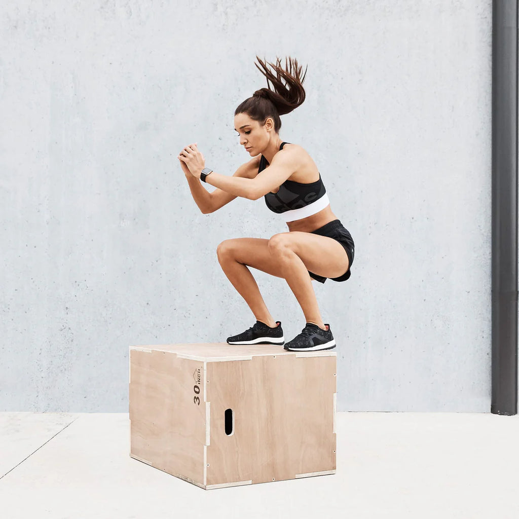 Woman performing box jumps on a wooden box against a light gray wall.