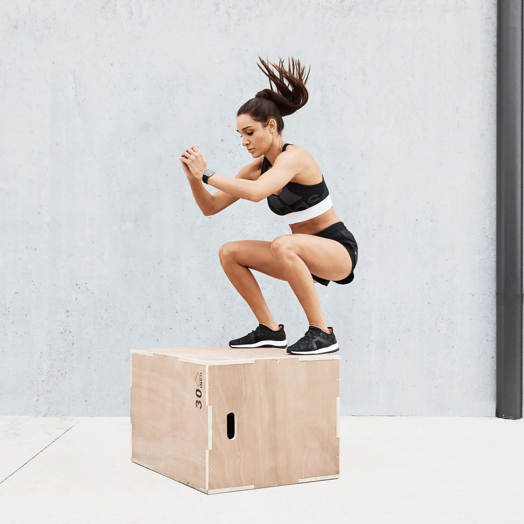 Woman performing box jumps on a wooden box against a light gray wall.
