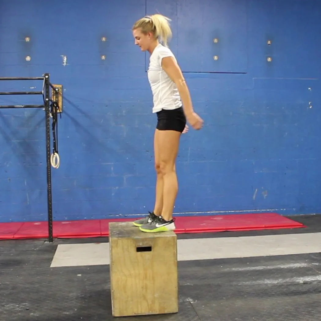 Woman performing box jumps on a wooden box in a gym setting.